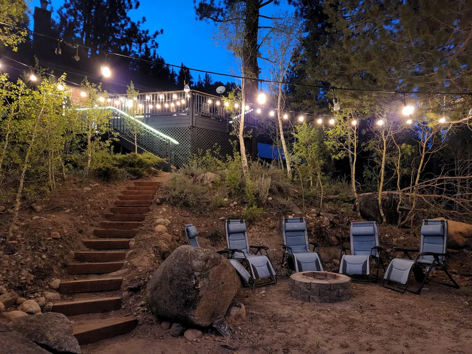 Outdoor chairs around the riverside fire pit beneath string lights, with stairs leading up to the cabin.