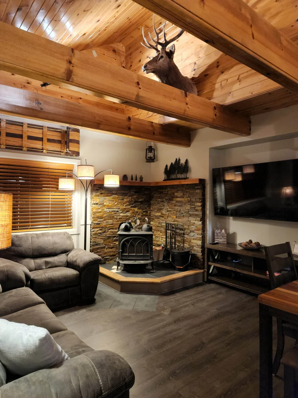 Cabin living room with gray seating, exposed wood beams, a stone hearth, wood stove and wall-mounted TV.