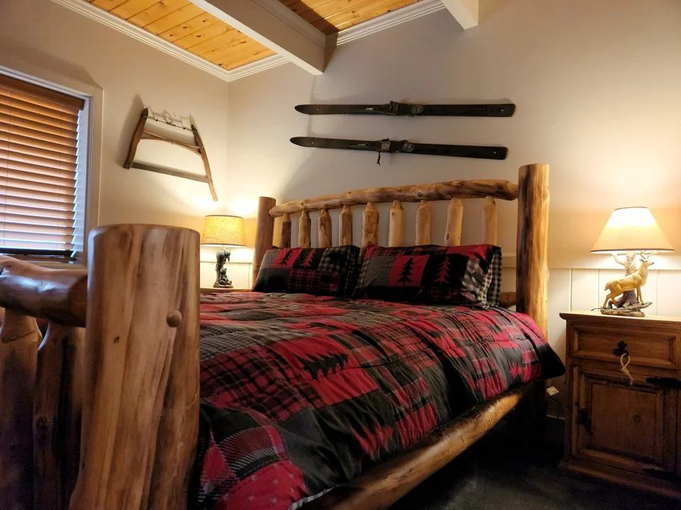 Cabin bedroom with a log-frame bed, red patterned bedding, bedside lamps and vintage skis on the wall.