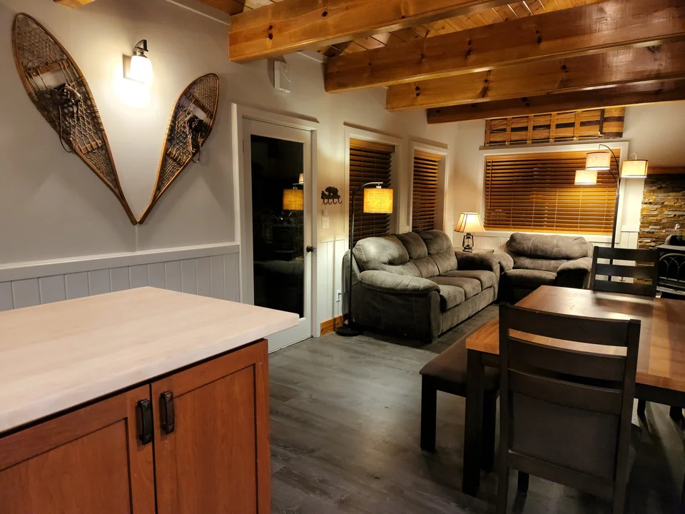 View from the kitchen counter toward the dining table and living room beneath exposed wood beams.